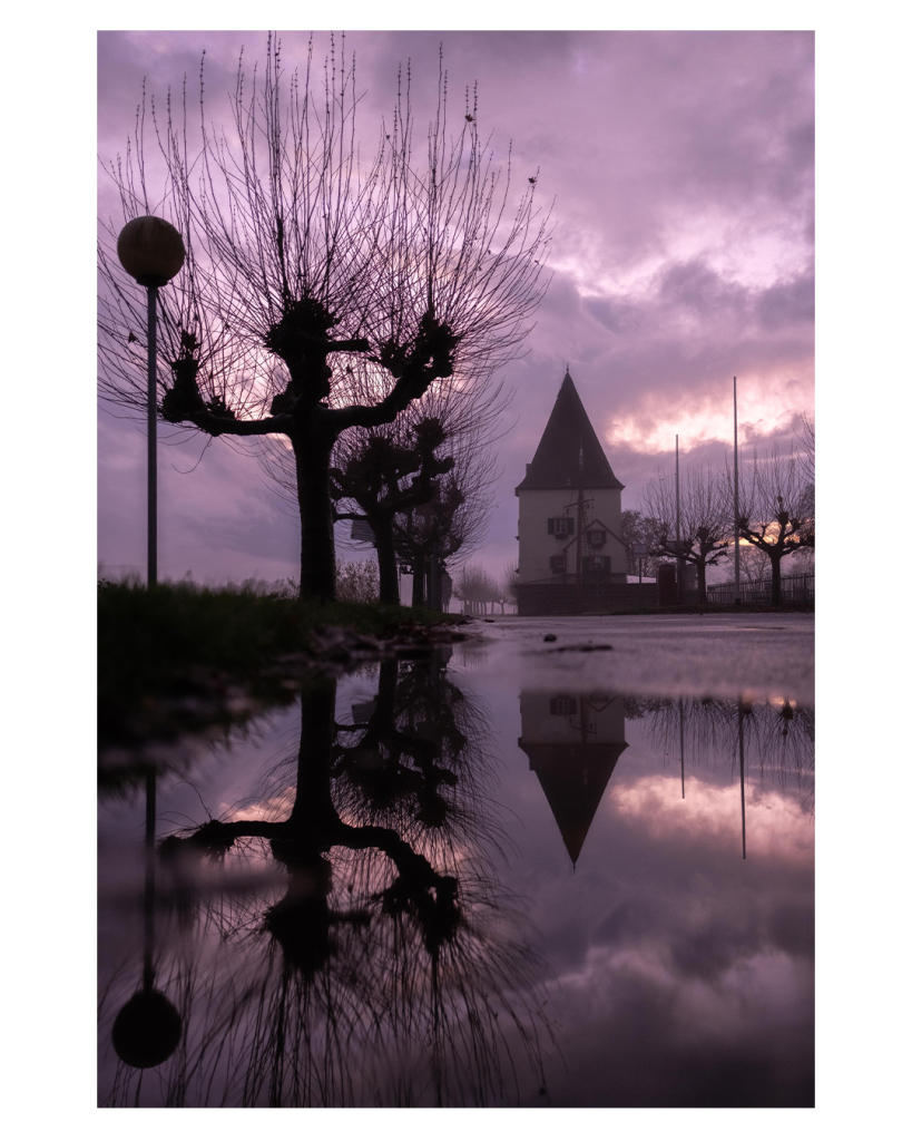 Foto im Hochformat. Nachts, neblig, Blick entlang eines Weges. In einiger Entfernung ist ein Gebäude mit Spitzdach. Davor steht der Mast eines Schiffes. Links sind mehrere Pappeln mit vielen knochigen einzelnen Äste. Daneben ist  eine einzelne Straßenlaterne. Die Kamera ist knapp über dem Boden über einer Pfütze. In der Pfütze spiegelt sich die Laterne, die Pappeln und der obere Teil des Gebäudes. Die allgemeine Stimmung des Bildes ist düster, der Himmel ist stark bewölkt und hinter den Wolken leuchtet es. 