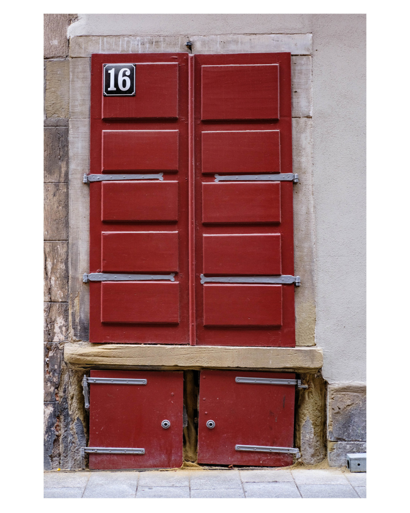 Foto im Hochformat. Eine rote Holztüre mit grauen Metallbeschlägen in einer alten Hauswand. Links oben prangt die Hausnummer 16, weiß auf einem schwarzen viereckigen Schild. 