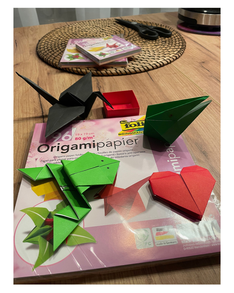 Foto im Hochformat. Auf einem Holztisch liegt eine Packung quadratisches farbiges Origami Faltpapier. Darauf liegen ein gefalteter grüner Frosch, ein schwarzer Schwan, ein rotes Herz und ein dunkelgrünes Schnabeltier (?). Dahinter liegt eine gefaltete rote Schachtel. Weiter hinten liegt weiteres Origamipapier. 