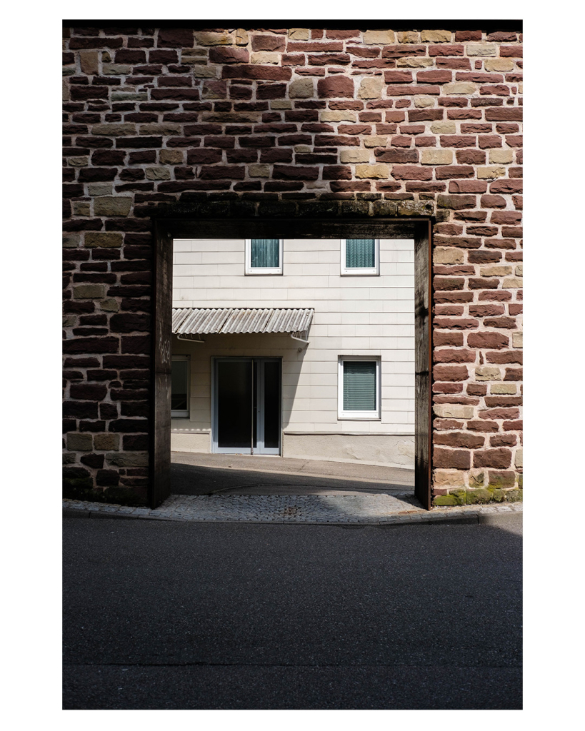 Foto im Hochformat. Blick auf eine alte bräunliche Steinmauer. Darin ist eine quadratische Öffnung. Dahinter ist eine Straße, dann ein weißes Haus. Drei Fenster, links eine Türe mit einem Wellblechvordach. 
