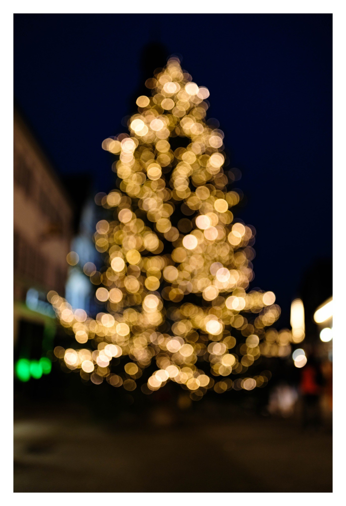 Foto im Hochformat. Nachts, das ganze Bild ist unscharf. Man erkennt einen riesigen Weihnachtsbaum mit elektrischen Kerzen. Durch die Unschärfe sind alle Lichter schöne, fast kreisrunde gelbe Lichtpunkte, sogenannte Bokeh-Highlights. 