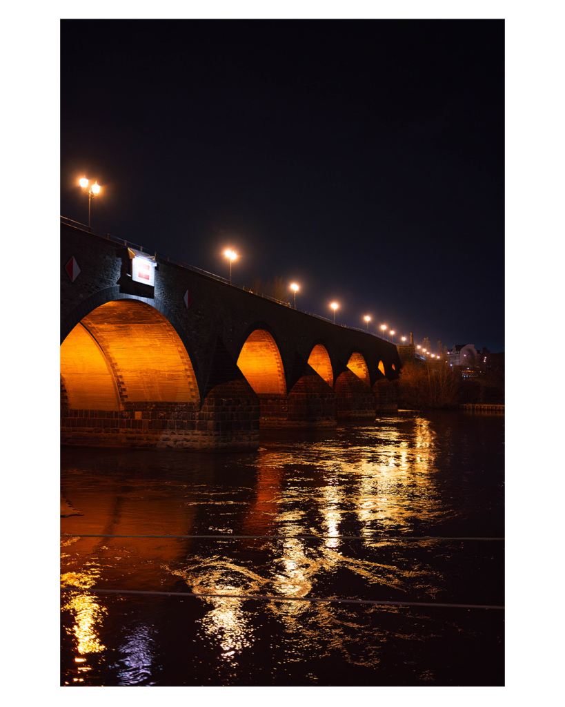 Foto im Hochformat. Es ist Nacht und Dunkel. Der Blick geht entlang einer steinernen Bogenbrücke. Die einzelnen Bögen sind beleuchtet. Auf der Brücke sind Straßenlaternen, die ebenfalls leuchten. Dies alles spiegelt sich im sich kräuselnden Wasser. 