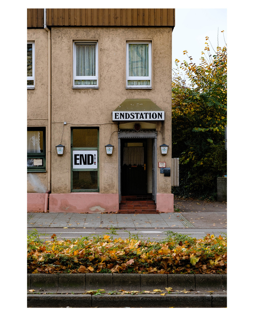 Foto im Hochformat. Blick über eine Straße, mittig ist ein Grünstreifen auf dem Herbstlaub liegt. Auf der anderen Straßenseite ist ein altes Gebäude. Darin ist eine Kneipe. Die Türe ist offen. Über der Türe an einem Vordach hängt ein Schild mit dem Namen. Endstation. Links am Fenster steh ebenfalls Endstation. Außerdem sind drei Schwaben-Bräu Lampen an der Wand.   
