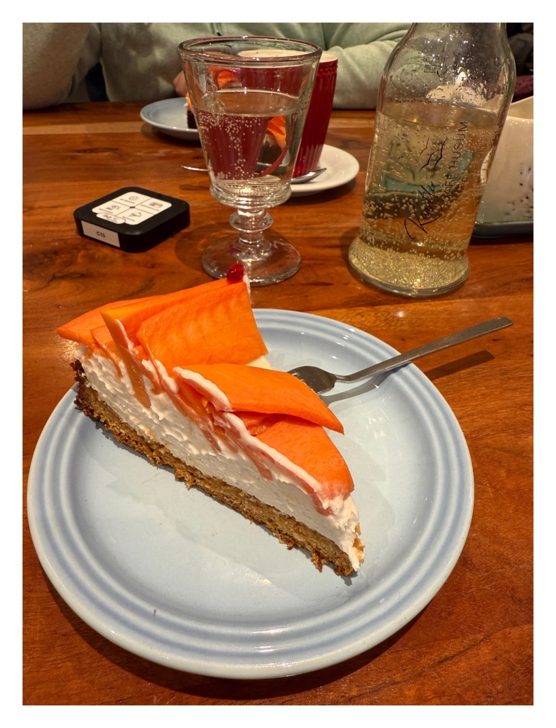 Foto im Hochformat. Auf einem Teller ist ein Stück vegane Papaya-Kokos-Tarte. Duenner hellbrauner Boden, eine dicke Schicht weiße Kokoscremé und darauf mehrere dünne Scheiben orangefarbene Papaya. Dahinter steht ein Glas und und Flasche mit Holunderlimonade. 