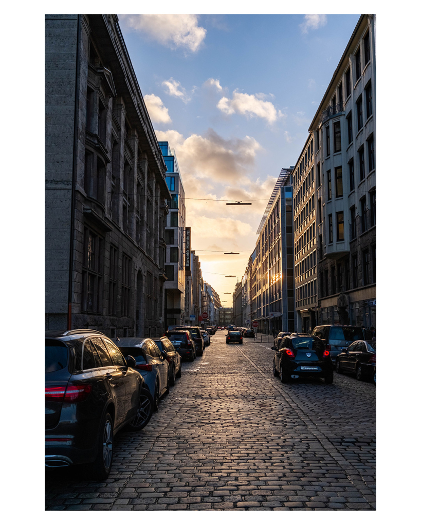 Foto im Hochformat. Blick entlang einer Straße in einer Großstadt. Links und rechts sind hohe Gebäude, teils älter, teils neuer, mit jeweils vielen Fenstern. Links und rechts parken Autos, auf der Straße aus Kopfsteinpflaster fahren auch welche. Der Himmel ist leicht bewölkt. Am Horizont sieht man die Sonne zwar nicht, man sieht aber ihr Licht. Sie geht bald unter und hüllt alles in ihren gelblichen Schein. 