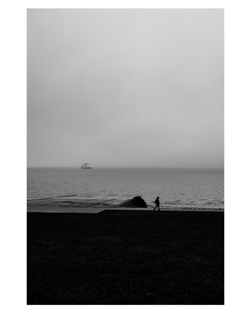 Schwarz-weiß Foto im Hochformat. Blickbaifs Meer. Es ist düster. Die Küste ist nur schwarze Silhouette. Am Meer läuft eine einzelne Person, siebter ebenfalls nur als Silhouette zu erkennen. Am Horizont fährt ein einziges Schiff. Der Himmel ist bedeckt und nimmt die Hälfte des Bildes ein. 