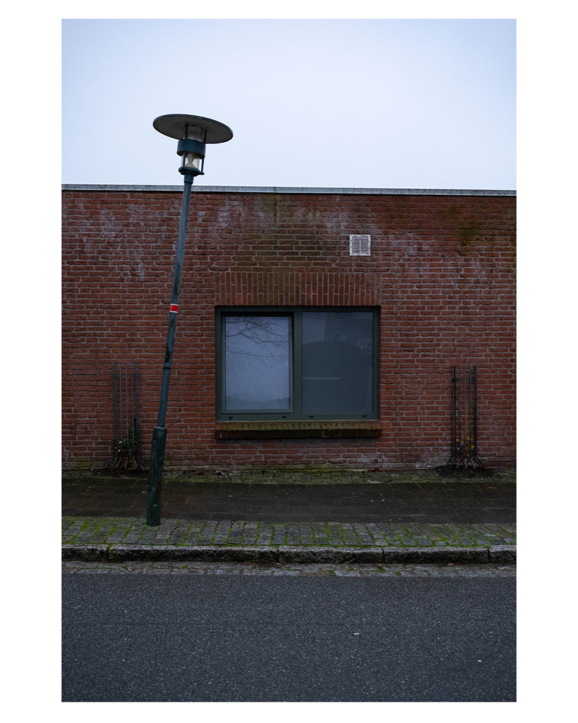 Foto im Hochformat. Blick über eine Straße auf ein rotes Gebäude aus Backstein. Mittig ist ein Doppelfenster, das linke ist gekippt. Über dem Fenster ist rechts ein weißes Blech mit Lüftungsschlitzen. Links und rechts des Fensters sind runde Metallgitter, in denen normalerweise Pflanzen wachsen. Vor dem Gebäude ist der Bürgersteig, teils mit Moos bewachsenem Kopfsteinpflaster, davor wiederum ist die Straße. Der Himmel über dem Gebäude ist bedeckt. Straße, Gebäude und Himmel nehmen jeweils ungefähr ein Drittel des Bildes ein, dadurch sieht alles sehr symmetrisch aus. Allerdings steht links vor dem Fenster eine schräge Straßenlaterne, da alles ins Ungleichgewicht bringt. Das Wetter ist nasstrüb. 