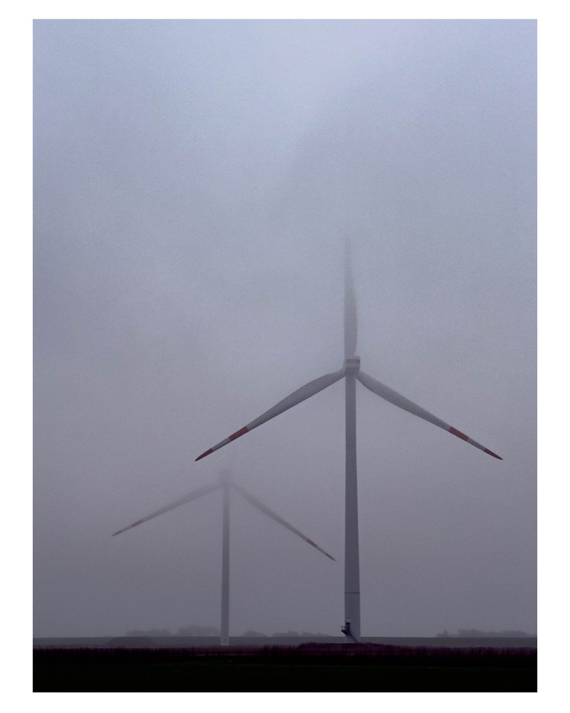 Foto im Hochformat. Zwei Windkraftanlagen im Nebel. Sie nehmen den großen Teil des Bildes ein, nur ein kleiner Streifen unten im Bild sind die Felder, auf denen sie stehen. Die Windräder stehen leicht versetzt hintereinander. Bei der vorderen verschwindet nur die obere Spitze im Nebel, die hintere ist nur halb zu sehen. Die jeweils drei Rotorblätter sind so ausgerichtet, das eins gerade nach oben zeigt. 