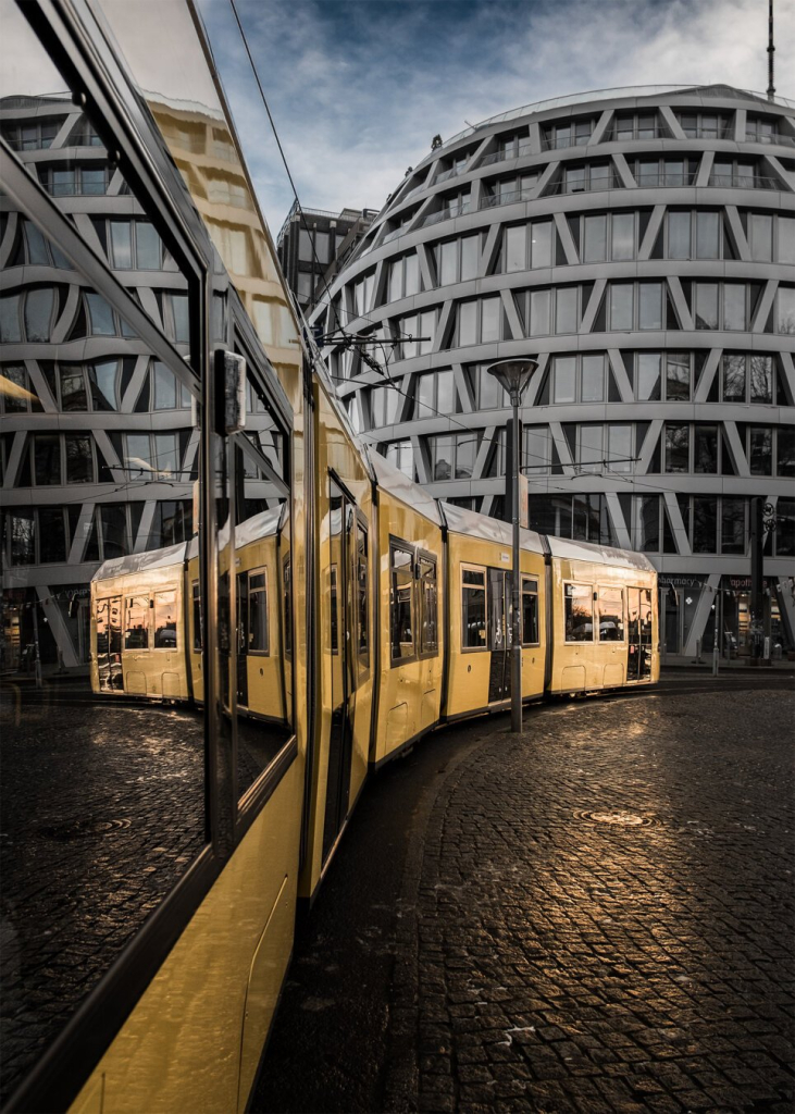 The image shows a yellow tram taking a curve on a cobblestone street. On the left, the tram reflects in its own windows, creating a symmetrical effect. In the background, a modern building with a distinctive white grid façade and large glass panes is visible. The sky is slightly cloudy, with shades of blue and gray. The light, likely from the sunset, casts warm reflections on the tram and the cobblestones. A streetlamp stands to the right of the tram.