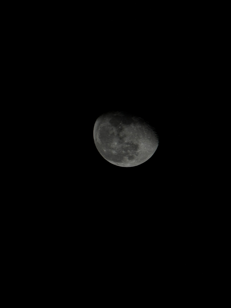 A pitch black photo, except for the moon in the center. It is a few days after the full moon, so it has begun to wane a bit. The details of the moons surface are visible, showing craters and lines. It’s a tad blurry and much darker than the other image. However, there is more detail visible because the brightness is lower.