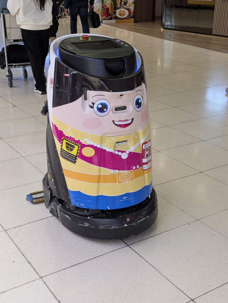 Photo of a roughly cylindrical, about 1 metre tall robotic floor cleaning machine. It has a colourful, cartoonish face.