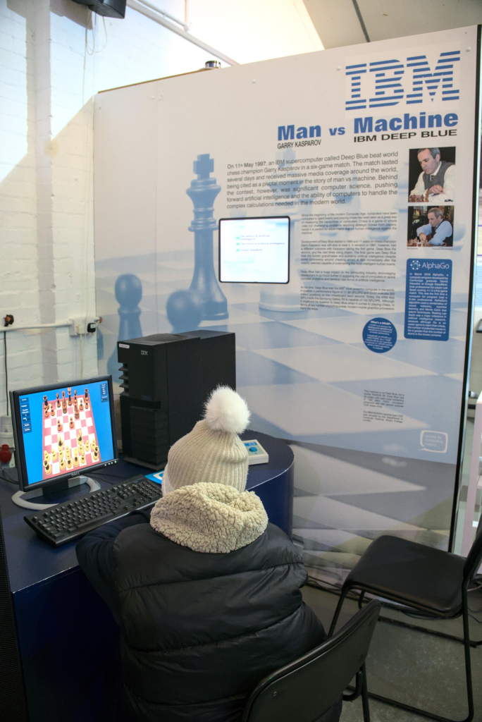 Photo of a museum display about the chess matches between IBM's Deep Blue and Gary Kasparov. In the foreground a child is playing chess against a computer.