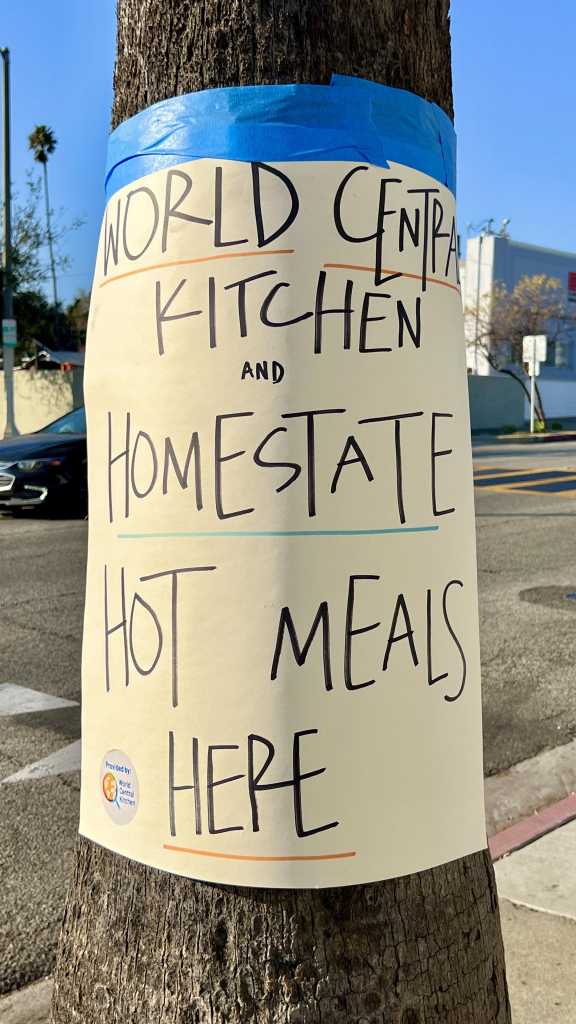 A sign affixed to a tree advertising "World Central Kitchen and Homestate Hot Meals Here." The sign features bold, handwritten text and is secured with blue tape. A small logo indicating the provision by World Central Kitchen is also visible at the bottom.