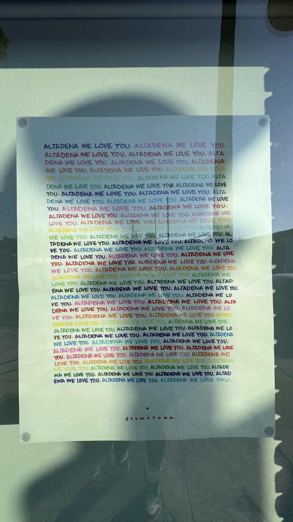 A colorful poster displays the phrase "Altadena We Love You" repeatedly in various hues. The bottom of the poster features the name "Stumptown." The poster is affixed to a wall with clear push pins.