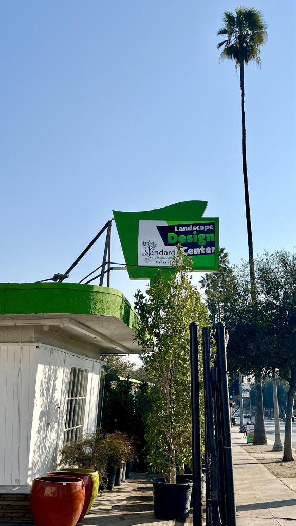 A landscape design center featuring a green and white sign. The building has a white exterior, ornamental plants, and colorful planters. A palm tree and a clear blue sky are visible in the background.