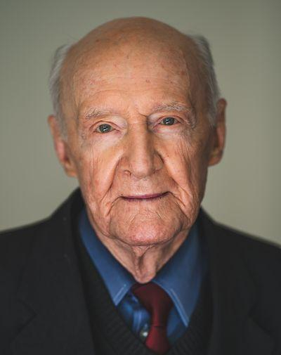 Close-up portrait of an elderly man with a solemn expression, wearing a dark suit and a red tie.