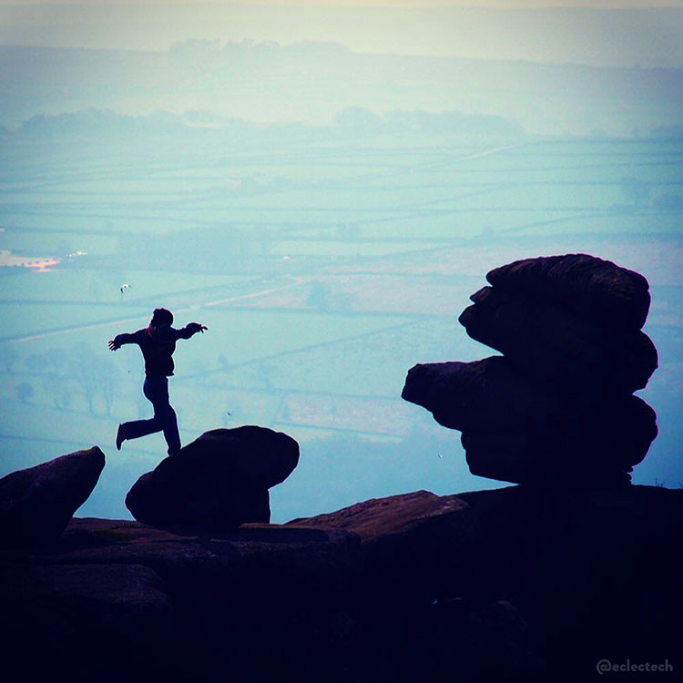 A square photo taken at Brimham Rocks in North Yorkshire, England. In the foreground are three large rocks, two smaller ones on the left and an interestingly layered large one on the right. There is a person caught mid-leap between the two smaller ones, arms outstretched to keep their balance. The rocks and person are all in darkness, and it looks like the land sharply drops away behind them with distant, hazy fields far below them in the distance, stretching to the top of the photo.