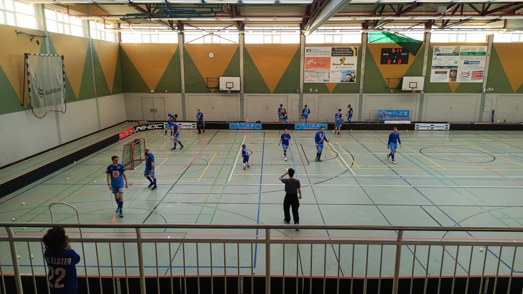 In einer Turnhalle bewegen sich Floorball SpielerInnen zum Aufwärmen im Spielfeld.
Am Geländer von den Tribünen schaut ein kleines Kind im Vereinstrikot dabei zu und sieht durch die Gitterstäbe vom Geländer