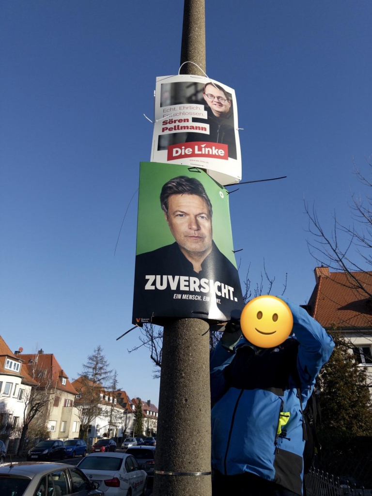 Eine Person steht auf einer Leiter und hängt ein grünes Plakat für Robert Habeck mit der Aufschrift “Zuversicht - Ein Mensch. Ein Wort.” auf. Das Gesicht der Person ist mit einem Smiley verdeckt. Über dem neu aufgehängten Plakat hängt bereits ein Plakat der Partei “Die Linke”. 