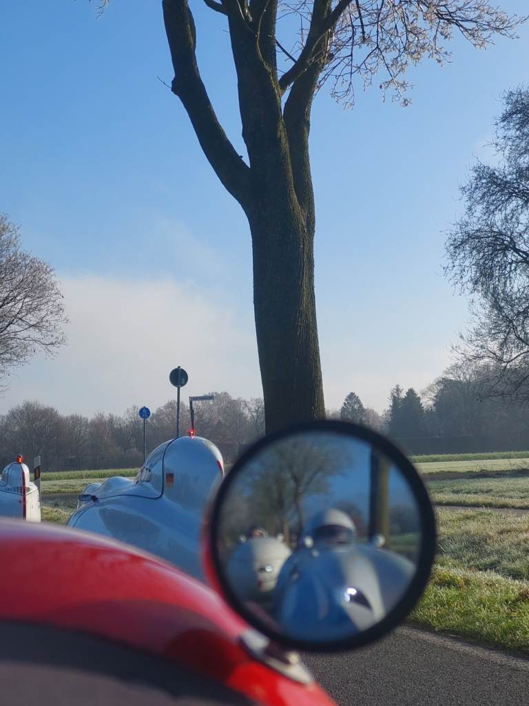 Blick über Rückspiegel und Kniebuckel meines roten Velomobils. davor zwei weiße Velomobilie (Milan), einer davon mit Deutschlandflagge aufgeklebt an der Kopfstütze. Im Spiegel sieht man unscharf noch zwei Velomobile von vorn, das hintere hat ein Gesicht mit herausgestreckter Zunge um die Lampen gemalt.  