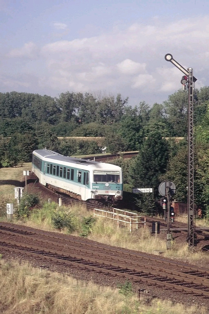 Das Bild zeigt einen mintgrün-weißen Dieseltriebwagen der Deutschen Bundesbahn, der aus einer Kurve auf einem leicht erhöhten Bahndamm in Richtung Kamera fährt. Im Vordergrund sind Gleise einer anderen Strecke zu sehen, die das Hauptmotiv kreuzen. Rechts steht ein mechanisches Formsignal, das auf "Fahrt" gestellt ist. Die Umgebung ist von dichtem Grün und Bäumen geprägt, der Himmel zeigt leichte Bewölkung an einem sonnigen Tag. Links und rechts der Strecke stehen kleine technische Einrichtungen und ein Zaun grenzt den Bahndamm ab. Ein Schild mit der Aufschrift „Düren W9“ ist erkennbar.

