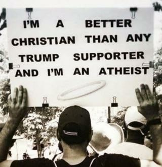 Black and white photo.
A man with his back to the camera holds up a sign that reads: "I'M A BETTER CHRISTIAN THAN ANY TRUMP SUPPORTER AND I'M AN ATHEIST."