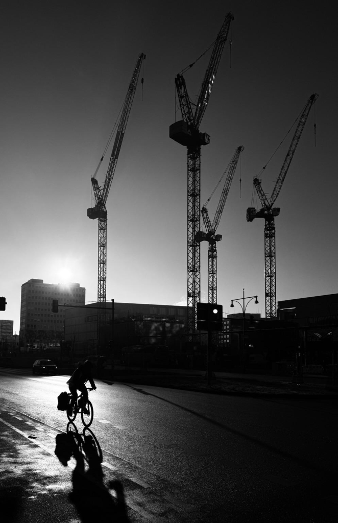 A silhouetted cyclist rides along a wet road with construction cranes in the background against a low sun, creating a dramatic black-and-white scene.