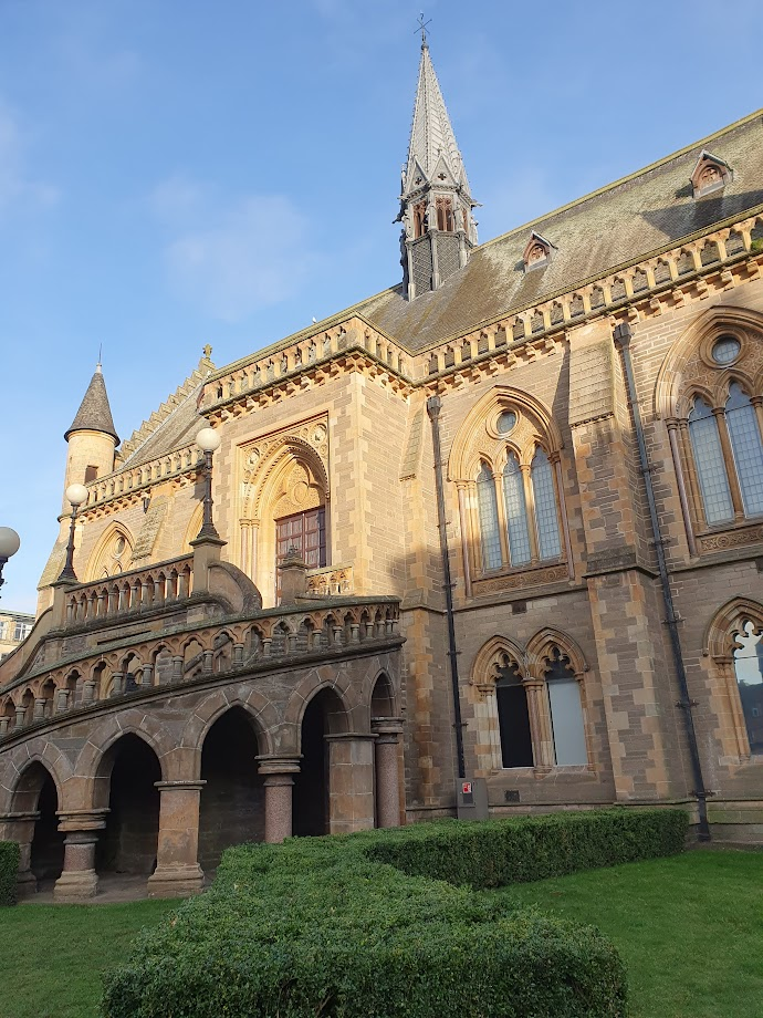 Photo. 
The McManus Galleries building in Dundee. A stone built Victorian Gothic style building. 
A winding staircase with arches underneath it leads to the first floor of the building. The building has large arched windows on ground and first floor. The stairs, external stair landing and around the roof of the building has a decorative balustrade, again made of stone with pillars and small arches. An ornate grey spire sits in the middle of the roof. 
There is lush green grass and a small hedge in front of the building, the sky is a lovely pale blue. 
In the photo, the ground floor and the grass are in the shade cast from buildings nearby, the first floor of the McManus and roof are lit by the sun. 