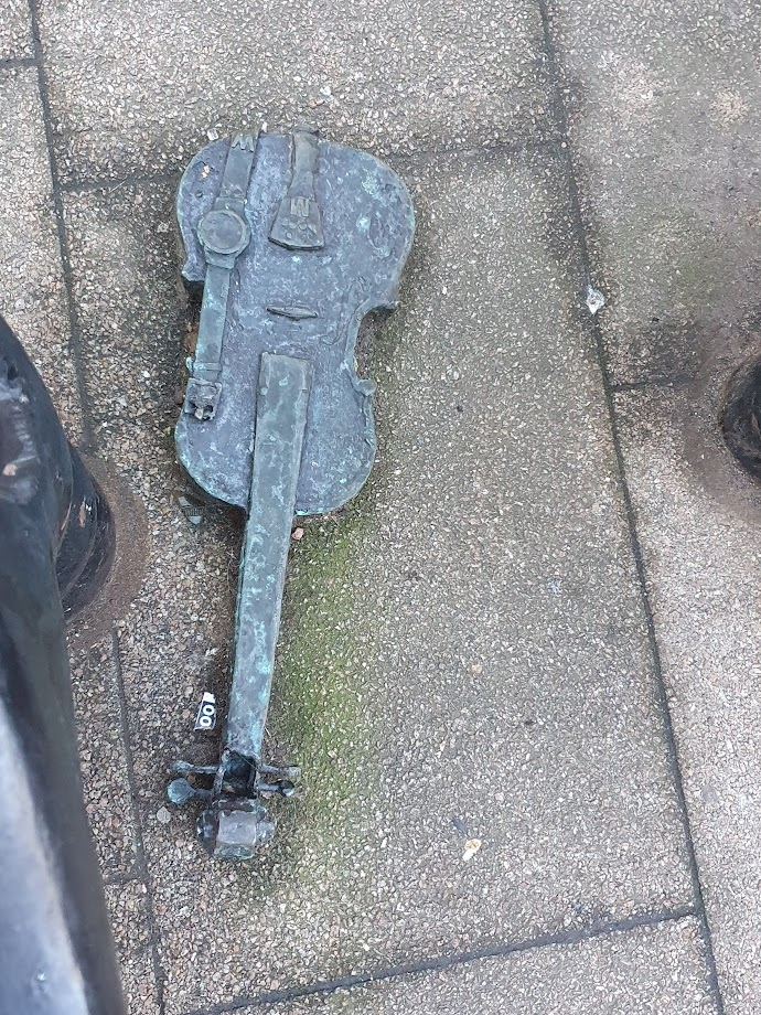 Photo. 
A small metal sculpture of a violin with a watch lying on it. The sculpture is on a paving slab and the metal feet of benches are on either side of the violin. 