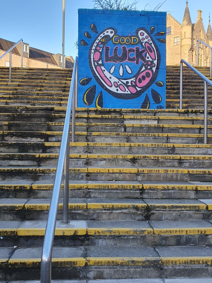 Photo. 
Concrete steps with the edges painted yellow, metal handrails at various points on the steps. What appears to be a random wall in the middle of the steps is actually used as a massive planter, steps continue at either side of it. 
The wall is covered in street art. The entire section of wall has a blue background, a large horseshoe is painted in white, yellow and pink. Yellow and black droplets surround the horseshoe. The words good luck are in the middle of the horseshoe. 
Buildings and blue sky can be seen beyond the back of the steps. 