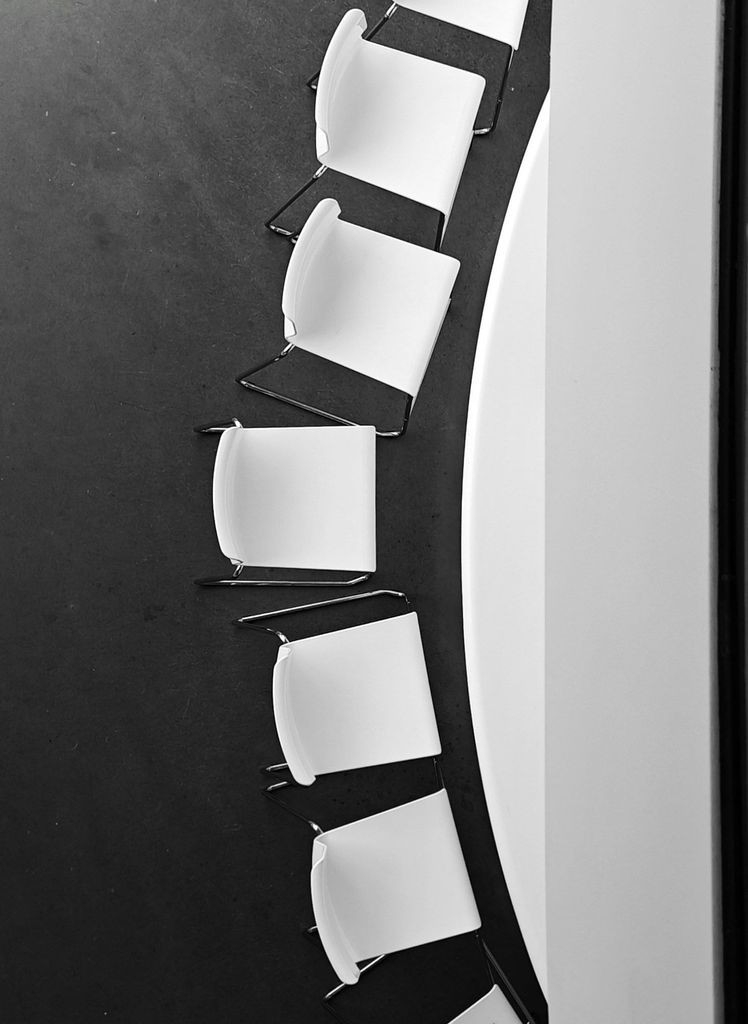 A black and white overhead photo of white modern chairs arranged in a curved line against a dark floor.