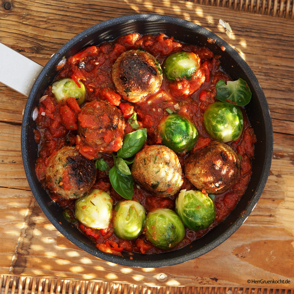 Heller Holzuntergrund. Darauf eine schwarze Pfanne. Darin: In Knoblauch gedünsteter Rosenkohl mit Dinkel-Bulgur-Frikadellen in einer leicht scharfen Tomatensauce mit italienischen Kräutern