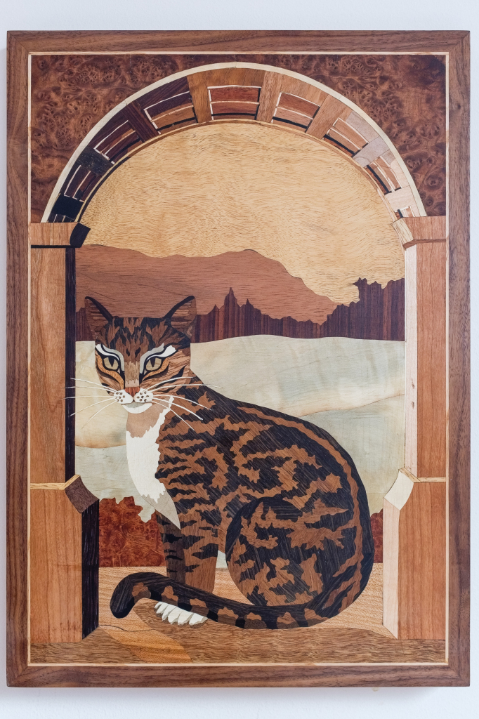 Photograph of a marquetry panel depicting a tabby cat seated below an arch, with a hilly vista in the background.