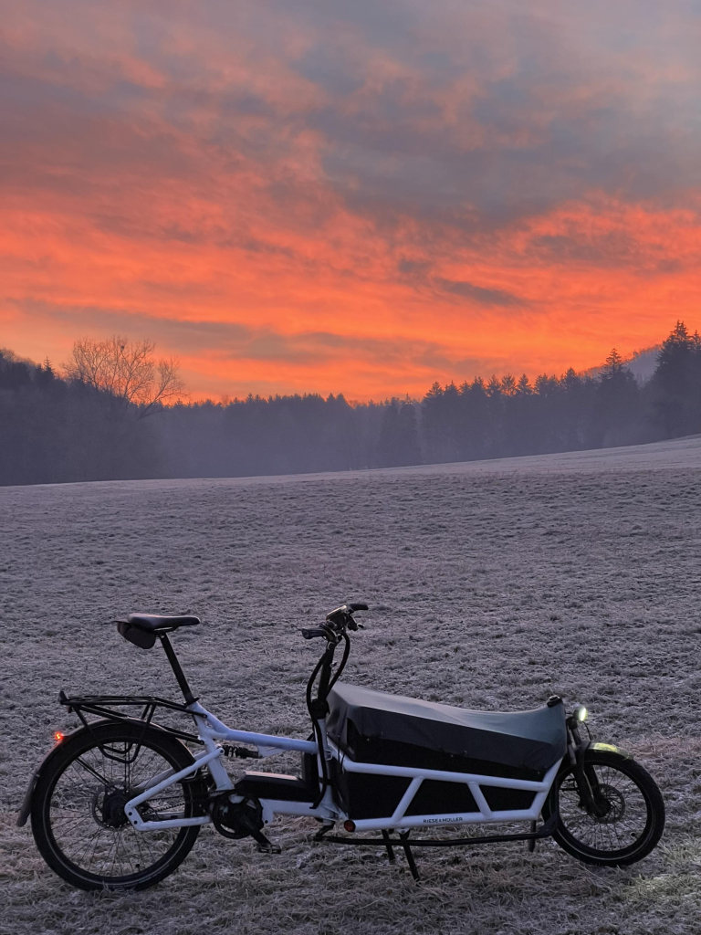 Ein weisses Lastenrad vor frostüberzogenen Feldern. Der Himmel ist bereits leicht Hellblau, am Horizont kündigt sich orange die aufgehende Sonne an