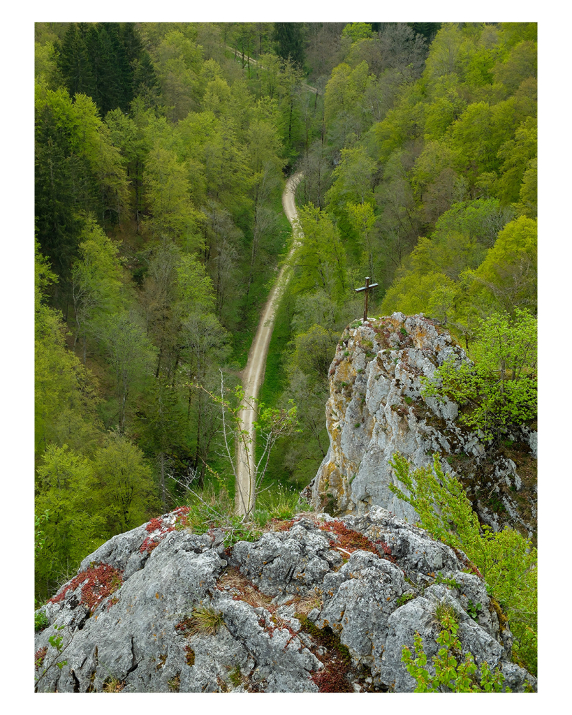 Foto im Hochformat. Blick von oben auf ein Tal in einem grünen Wald. Durchs Tal schlängelt sich ein hellbrauner Weg, von unten nach oben. Im Vordergrund ist hellgrauer Felsen, von dem aus das Foto gemacht wurde. Weiter unten ist ein weiterer Felsen, auf diesem steht ein Kreuz. 