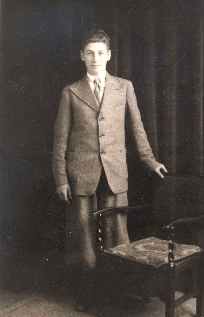 A vintage black and white photo of a boy standing next to a chair, dressed in a three-piece suit with a patterned tie.