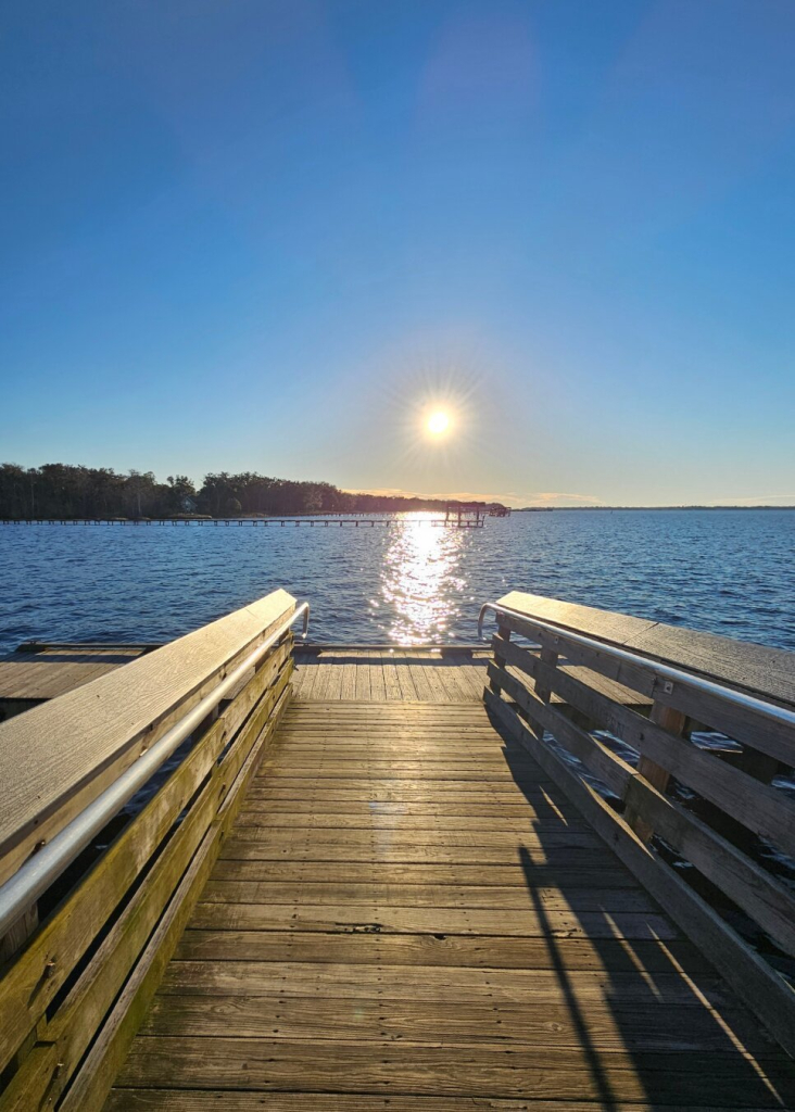 From "County Dock" off Mandarin Road in Jacksonville, Florida,  view from the gangway over the blue Saint Johns River, beneath a big blue Florida sky,  with a starburst sun in the center as it lowers in the sky approaching the horizon far in the distance.