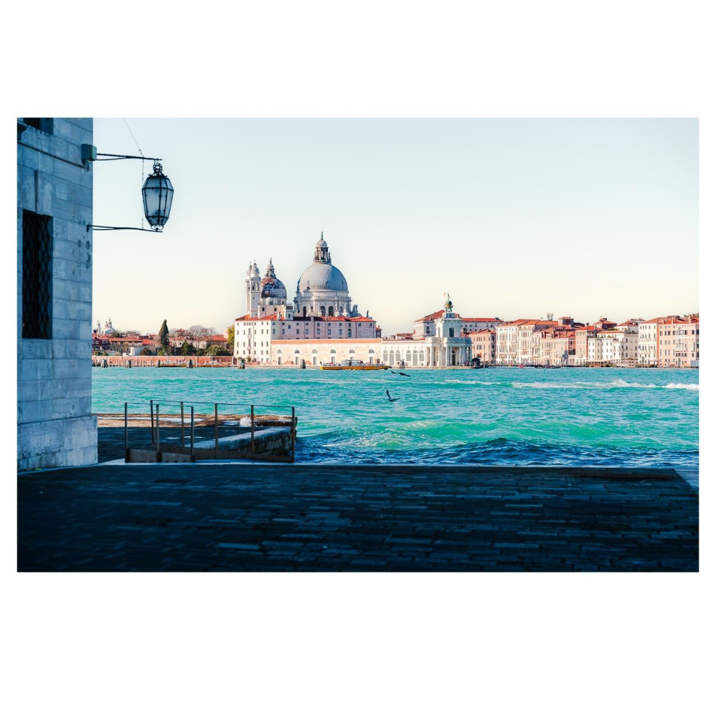 Szene in Venedig. Im Vordergrund ein Gebäude mit künstlich angeleger Promenade. Dann blicken wir ca. 700m über die Lagune. Im Hintergrund die Silhouette der Stadt. 