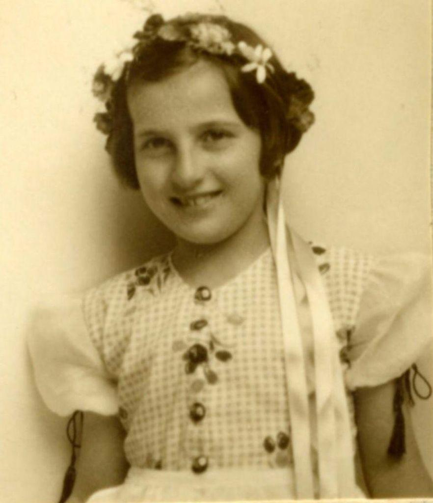 Vintage portrait of a young girl wearing a dress adorned with flowers and ribbons, smiling at the camera.