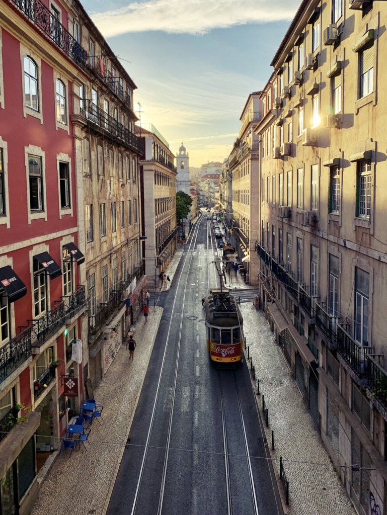 A street car coasts down a street with golden sunlight bouncing off the buildings on the right side of frame to illuminate the buildings on the left side of frame.