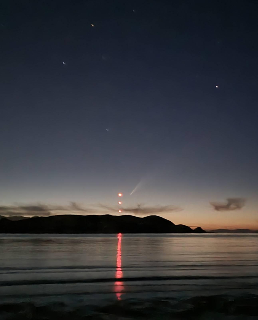 Calm bay, reflecting red pilon lights, sun just gone down on a clear warm summer's evening, and the comet can be seen wooshing away.