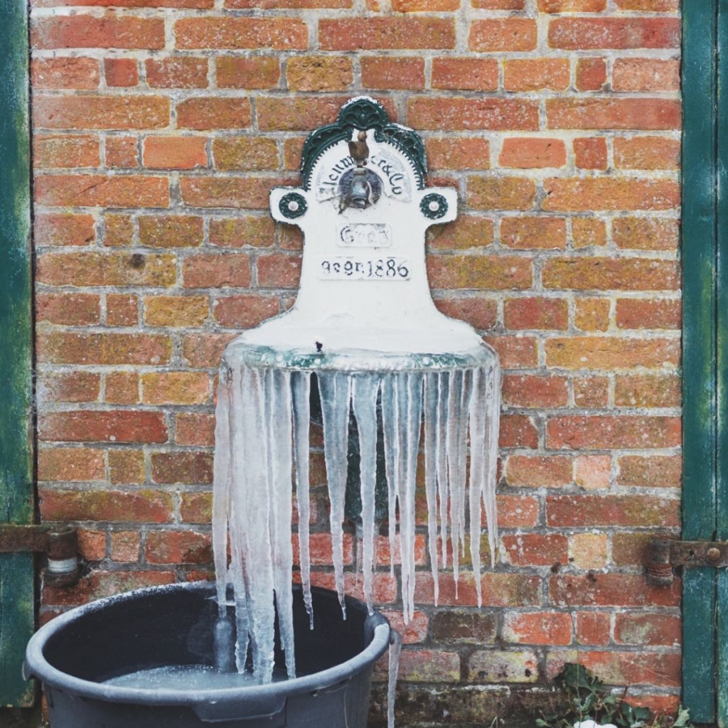 Eine historische weiße Tränke von 1886 an einer roten Backsteinmauer eines Schuppens ist über Nacht übergelaufen. Dank eisiger Temperaturen ist die Tränke nun gesäumt von Eiszapfen, die wie kleine erstarrte Wasserfälle aussehen.