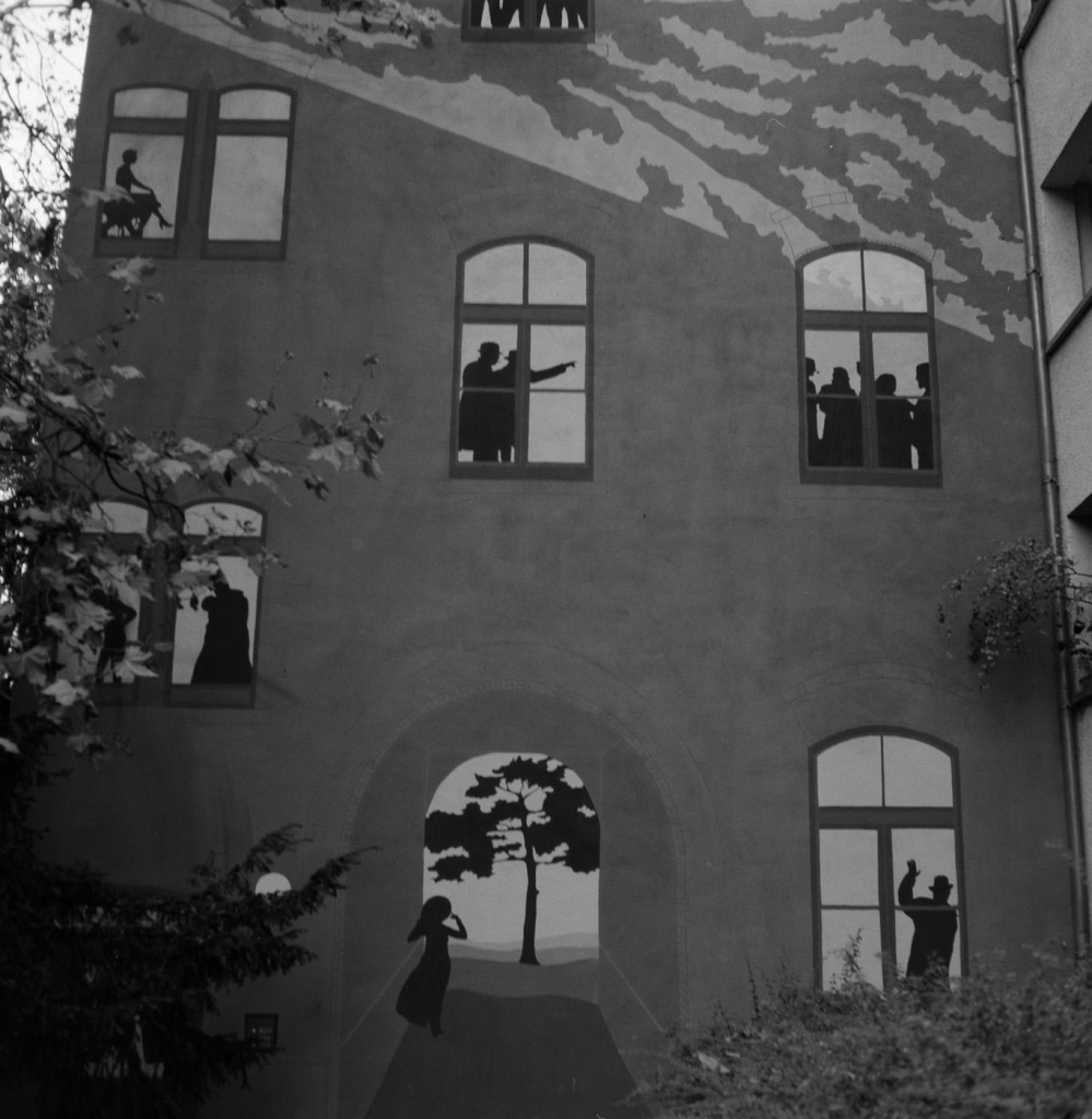 Eine Hauswand in einem Hinterhof der Kölner Südstadt. Auf die Wand sind Fenster gemalt, in denen Menschen zu sehen sind. Unten ein aufgemalter Durchgang mit einem Baum dahinter. Ein Kind läuft dadurch. 

Das Foto ist in schwarzweiss. 