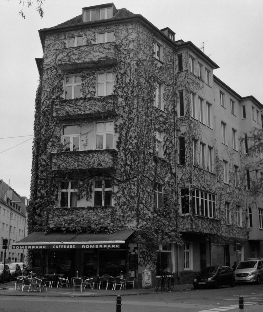 Ein mit Efeu bewachsenes, mehrstöckiges Eckhaus. Das Gebäude wird nach vorne immer schmaler. Unten ist das Cafehaus Römerpark, mit Stühlen draussen und einem grossen faltbaren Dach darüber. Das Foto ist in schwarzweiss und auf Film entstanden.