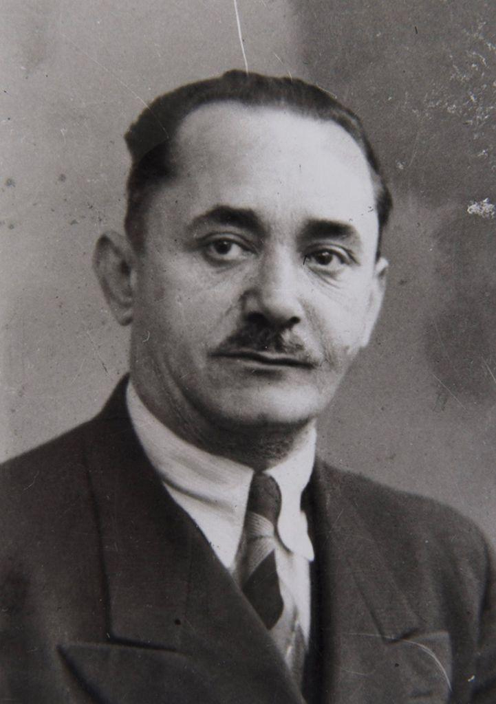 Black-and-white portrait of a mature man, featuring a person in a suit with a neat tie and a distinct mustache, looking directly at the camera.
