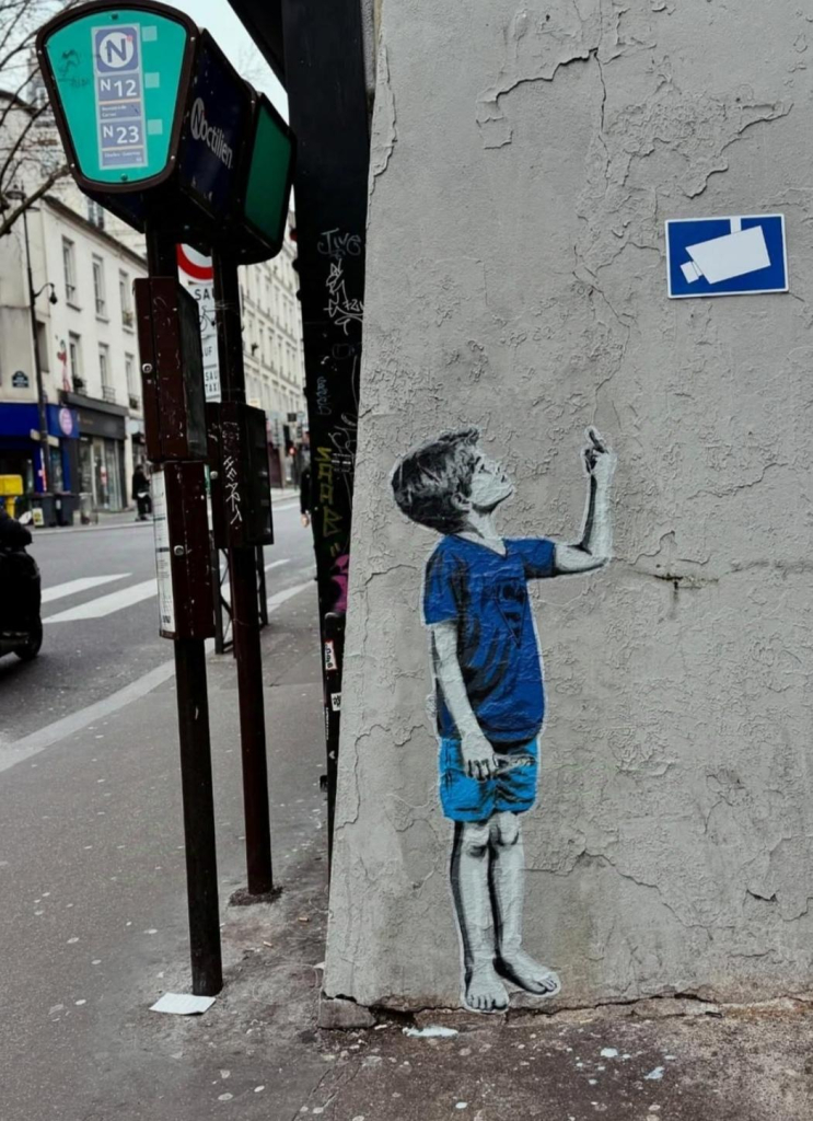 Streetart. A small self-painted picture of a little boy has been placed on the corner of a house under a blue sign indicating camera surveillance. The boy is dressed in a blue T-shirt and shorts, looking up at the camera pictogram and showing his middle finger. (A bus stop and the street can be seen next to the mural)