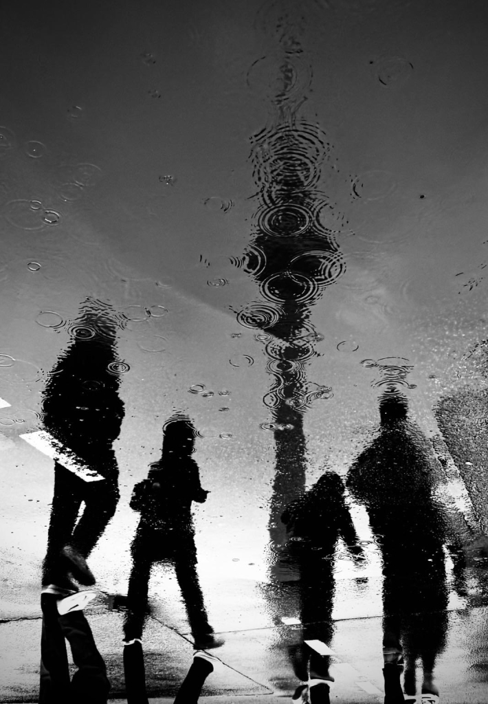 A black and white reflection in a puddle shows the silhouettes of people walking, with ripples of raindrops visible on the water's surface. The image captures an urban scene, likely with a tower visible in the reflection above.