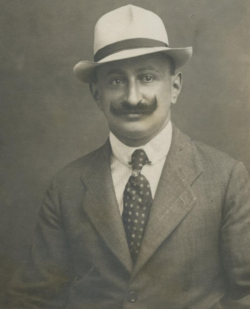 Vintage black-and-white portrait of a man wearing a hat, a three-piece suit, and a polka-dot tie, exuding a confident smile.