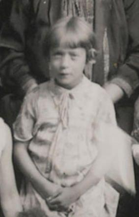 A vintage black and white photo of girl, wearing a light-colored dress and sitting with hands clasped together.