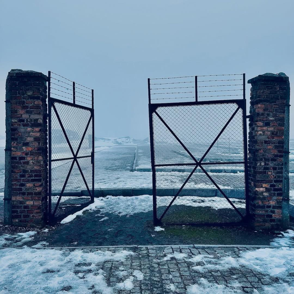 The image depicts a set of rusted metal gates attached to two weathered brick pillars, standing open against a bleak, snow-covered landscape. The ground and surrounding area are blanketed in snow, and the horizon is obscured by dense fog, creating a cold and somber atmosphere. 